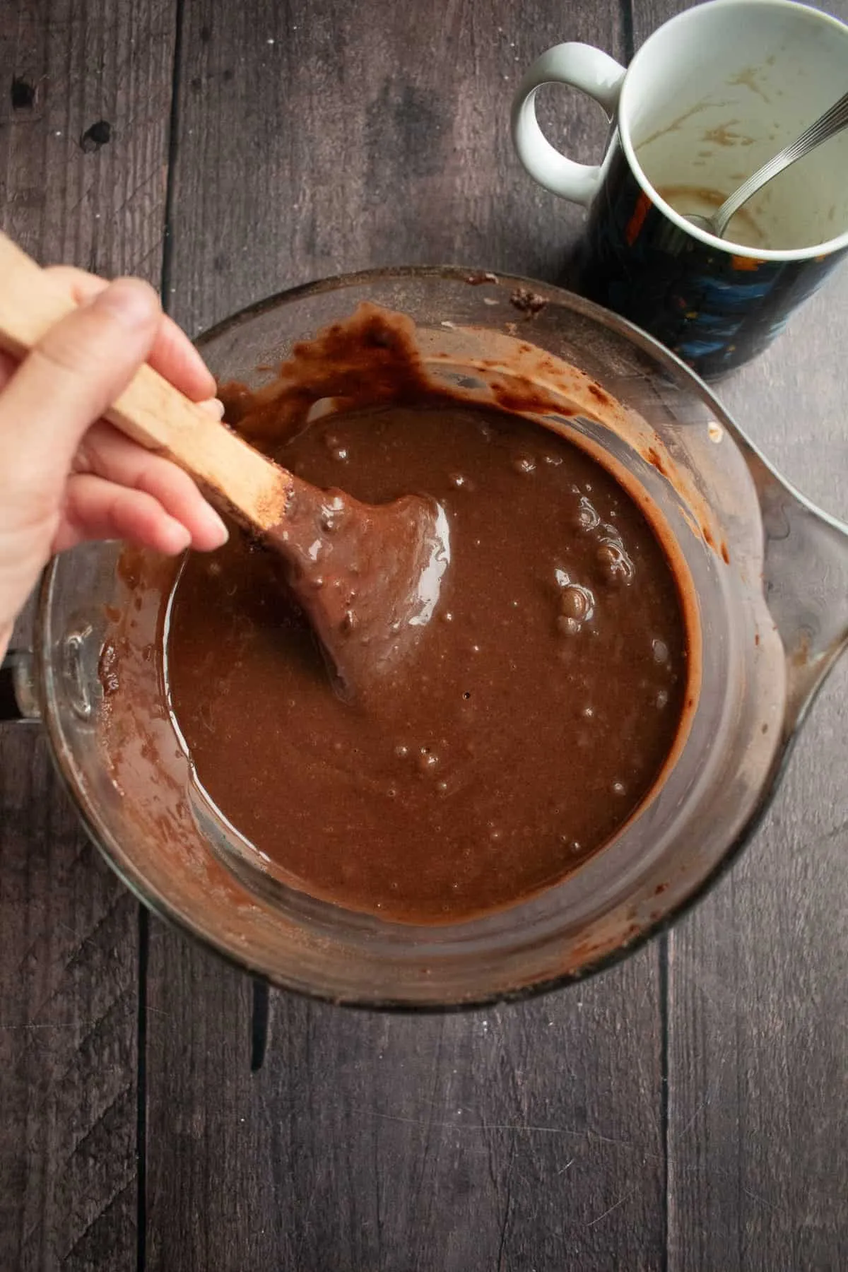 Eine Hand rührt glatten Schokoladenkuchenteig in einer Rührschüssel neben einer leeren Kaffeetasse.