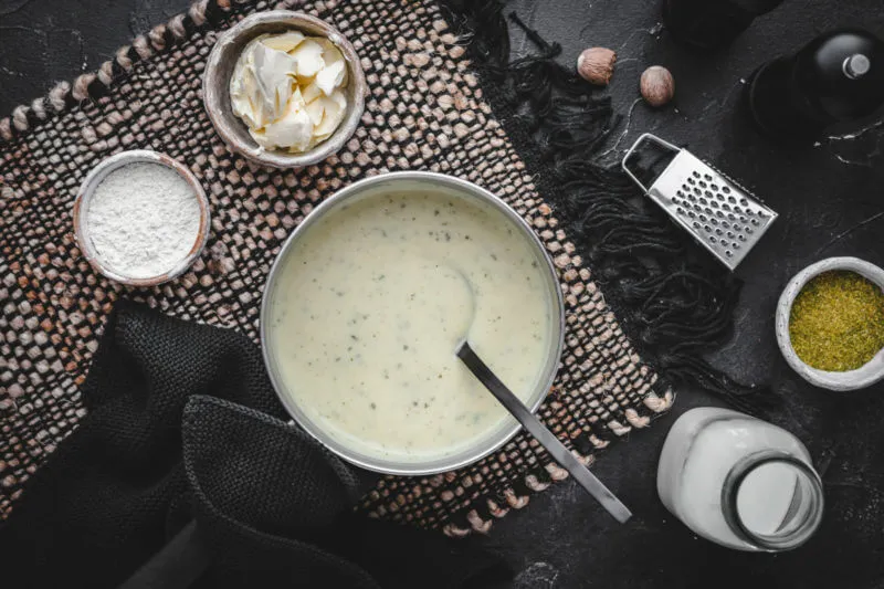 Eine helle Schüssel gefüllt mit fertiger Bechamelsoße - drumherum sind alle Zutaten für die Soße in kleinen Gefäßen angeordnet, darunter Butter, Mehl, Milch und Muskatnuss.