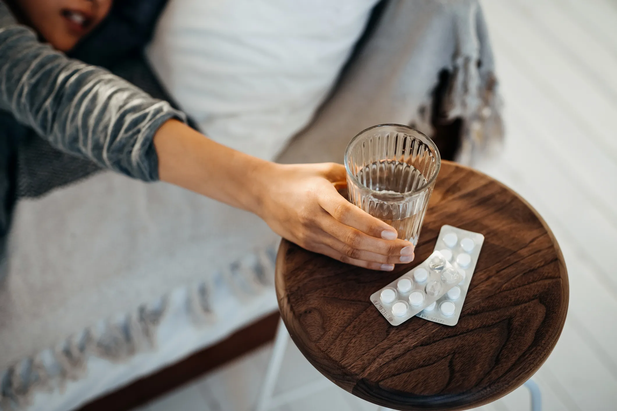 Eine junge Asiatin liegt krank im Bett und greift nach einem Glas Wasser und Medikamenten auf dem Nachttisch.