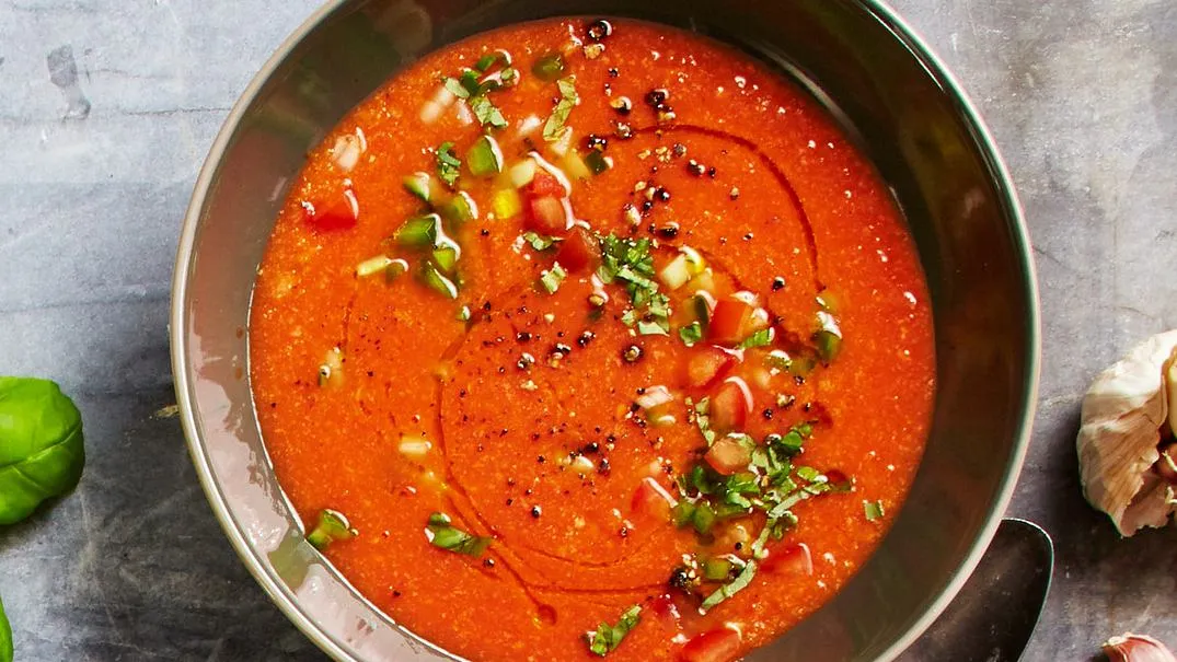 Eine klassische, kalte Gazpacho in einer Schüssel, garniert mit frischen Kräutern und einem Schuss Olivenöl, bereit zum Servieren an einem heißen Tag.