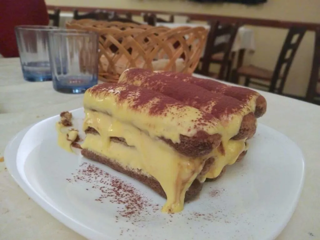 Eine klassische Scheibe Tiramisù auf einem weißen Teller, dessen Schichten aus kaffeeträchtigen Biskuit und reichhaltigem, cremigem Mascarpone mit Kakaopulver bestäubt sind, ein typisch italienisches Dessert aus Rom.