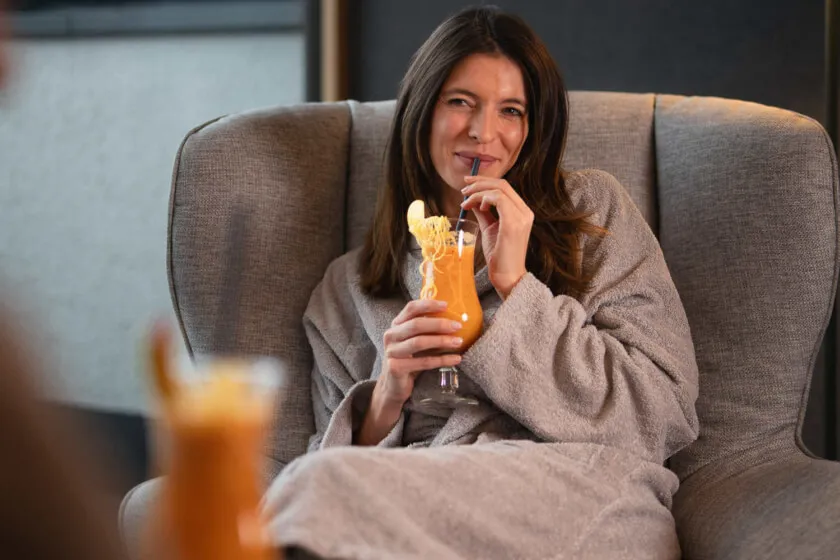 Eine lächelnde Frau im Bademantel entspannt in einem Sessel und trinkt einen fruchtigen Cocktail im Wellnessbistro.