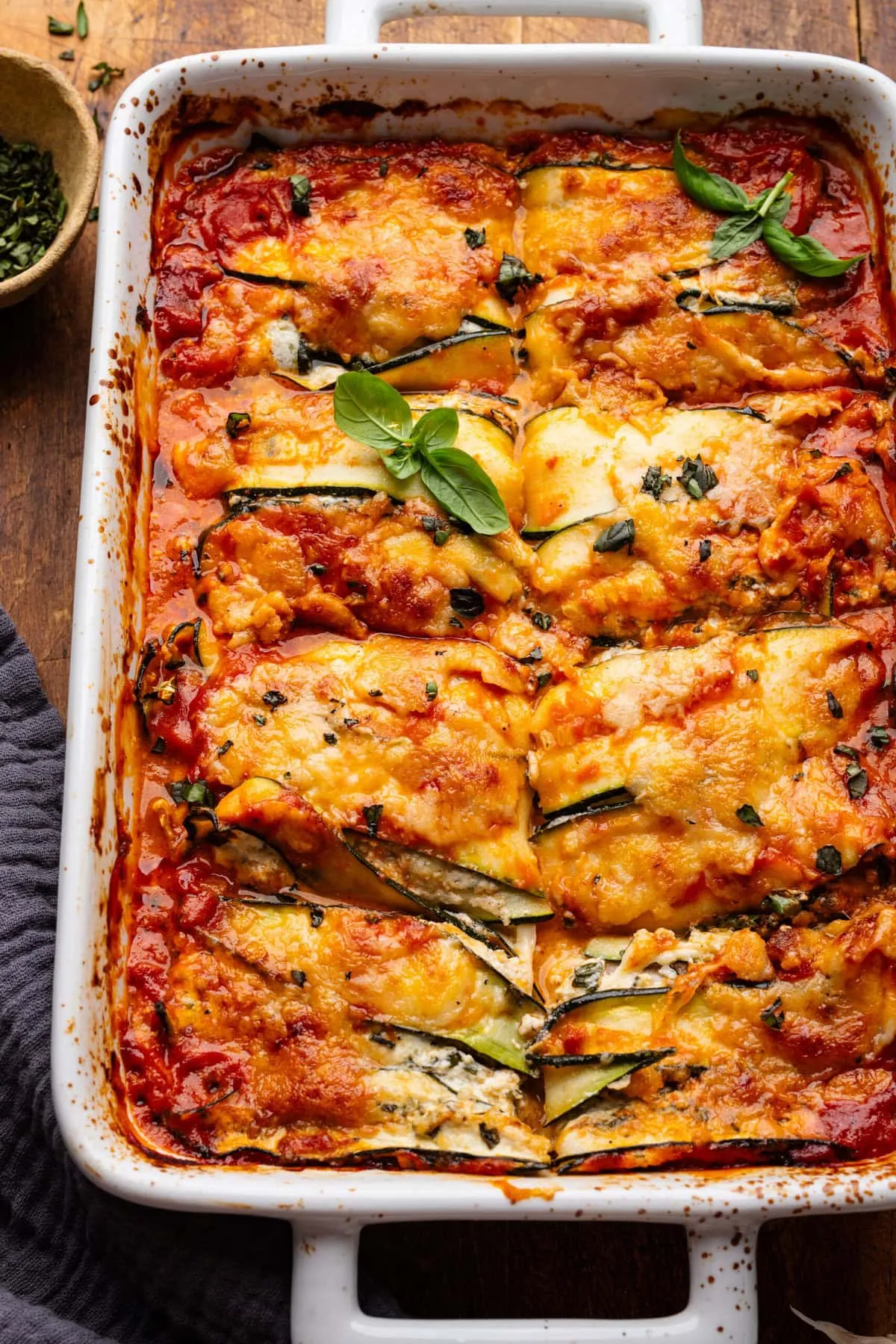 Eine rechteckige weiße Auflaufform mit Zucchini Lasagne, in acht Stücke geschnitten. Die Form steht auf einem Holzbrett, dekoriert mit frischen Kräutern und einer dunklen grauen Serviette.