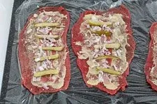 Eine Rinderroulade wird mit Senf bestrichen, Speck, Gewürzgurken und Schalotten belegt, bereit zum Einrollen.
