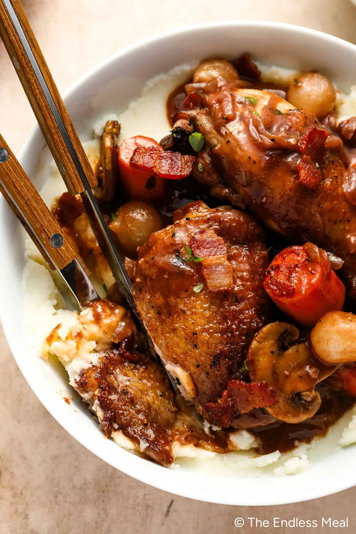 Eine Schüssel mit klassischem Coq au Vin mit zartem Hähnchen, Karotten, Pilzen und Zwiebeln auf cremigem Kartoffelpüree. Zwei Holzbestecke ruhen einladend in der Schüssel.