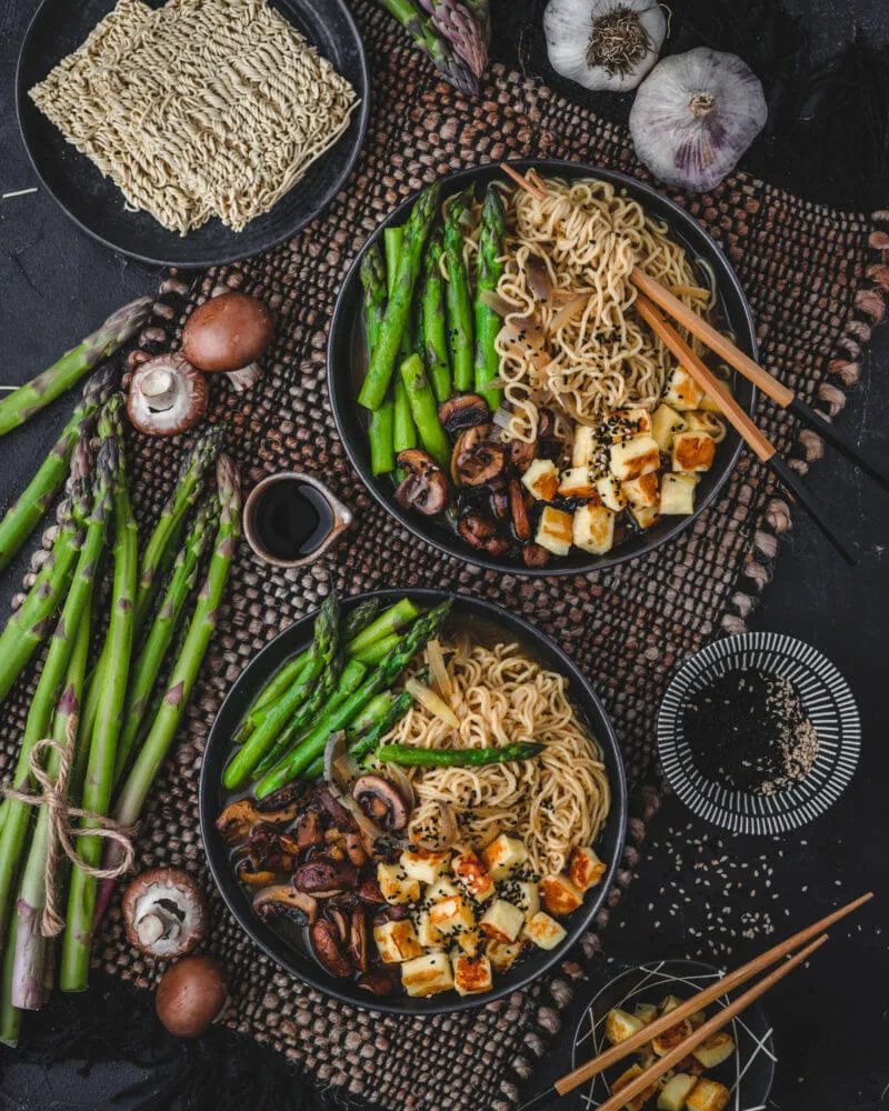 Eine tief dargereichte vegetarische Ramen Suppe mit goldbraun gebratenem Halloumi, frischem grünem Spargel und saftigen Pilzen, elegant angerichtet
