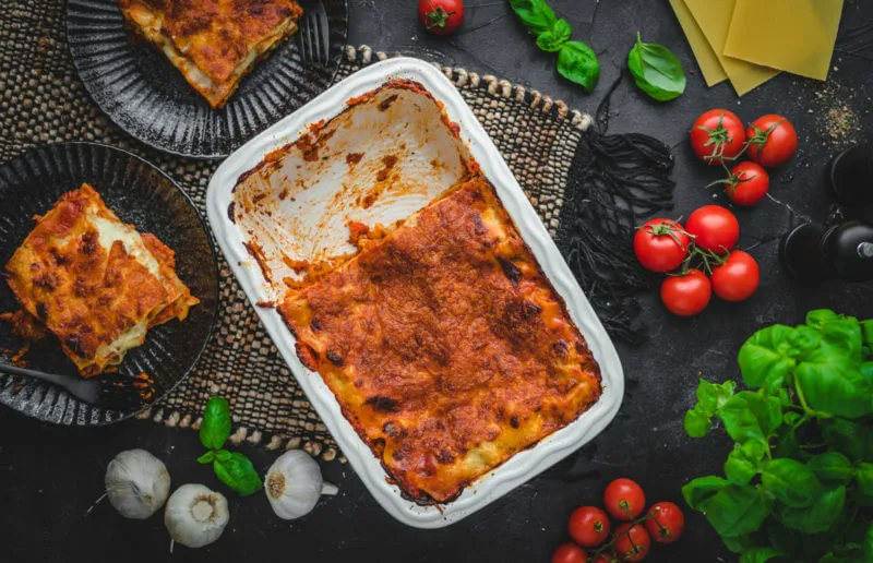 Eine weiße Auflaufform gefüllt mit vegetarischer Lasagne auf dunklem Untergrund. Daneben Garnitur und zwei schwarze Teller mit jeweils einem Stück Lasagne.