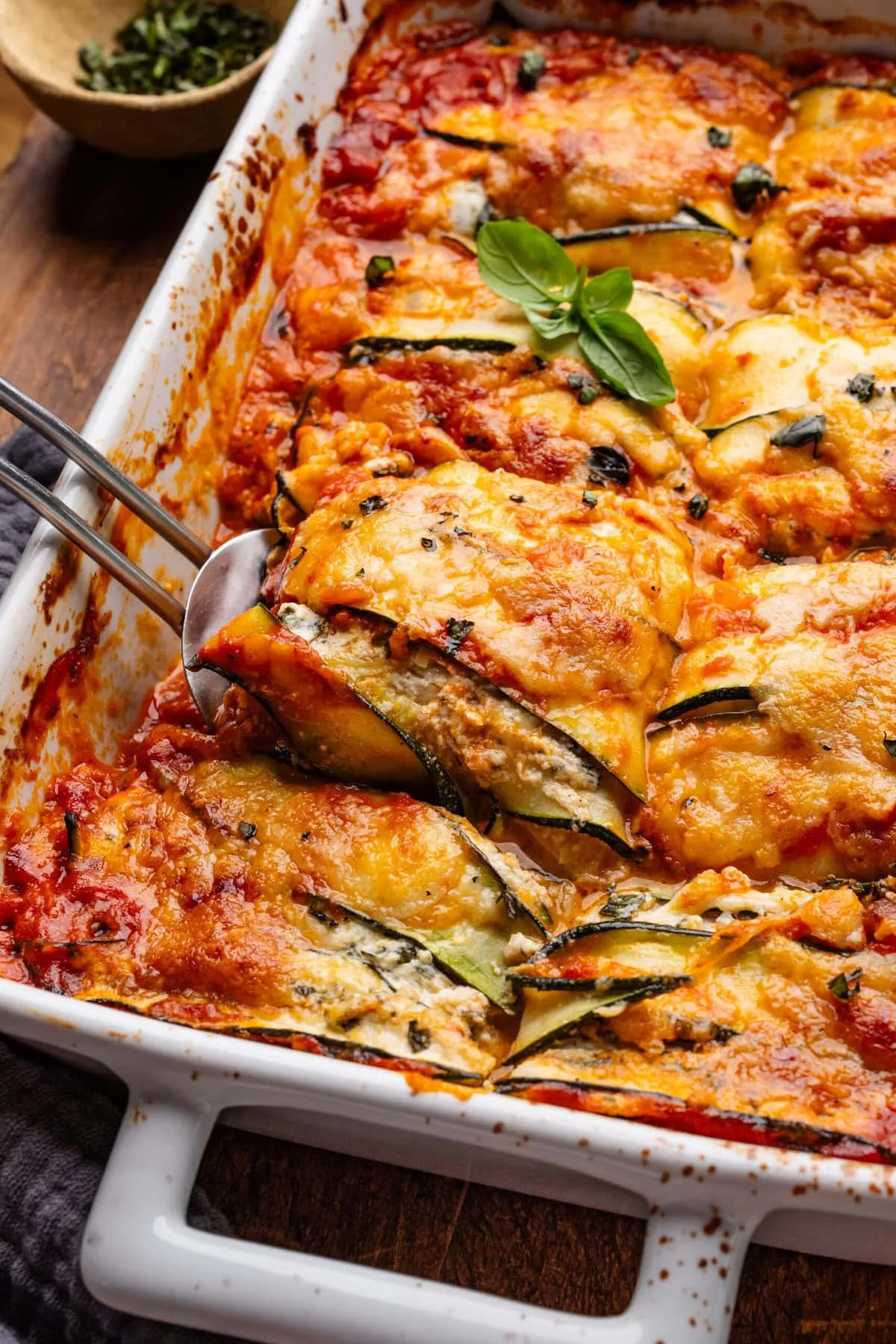 Eine weiße Auflaufform mit Zucchini Lasagne, ein Stück wird mit einem Spatel herausgehoben, um die Schichten zu zeigen.
