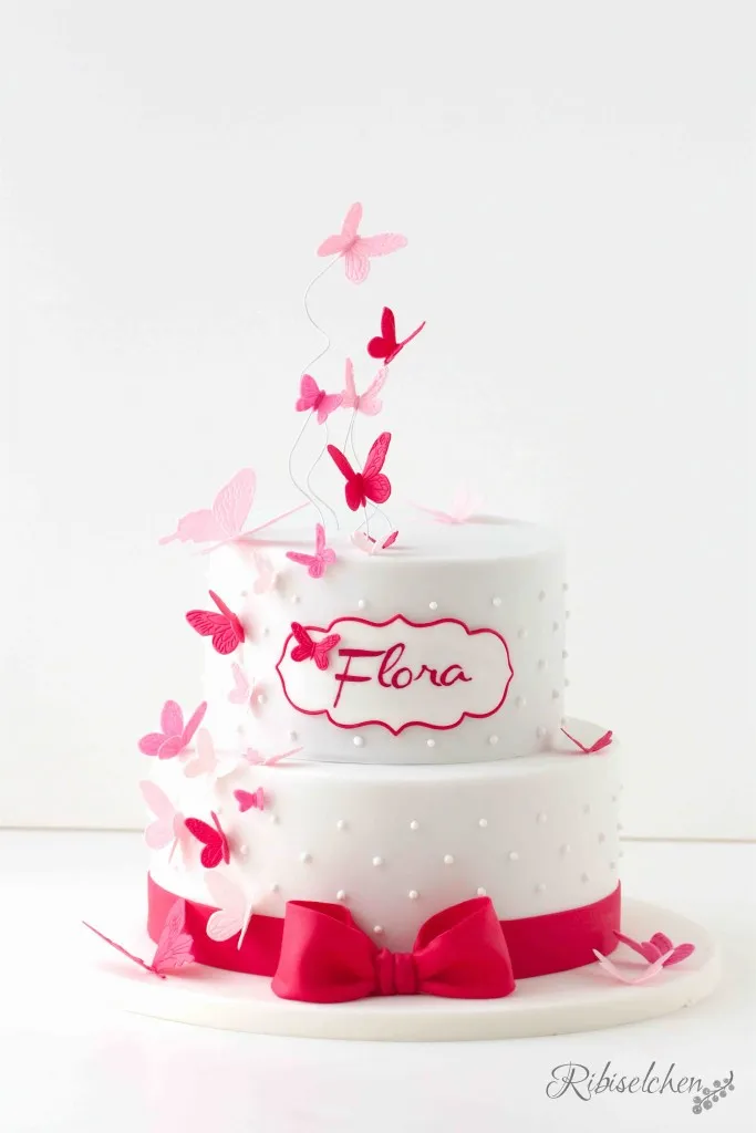 Eine wunderschöne Tauftorte in Rosa und Pink, verziert mit zarten Schmetterlingen, Royal Icing Punkten und einer eleganten Fondantmasche