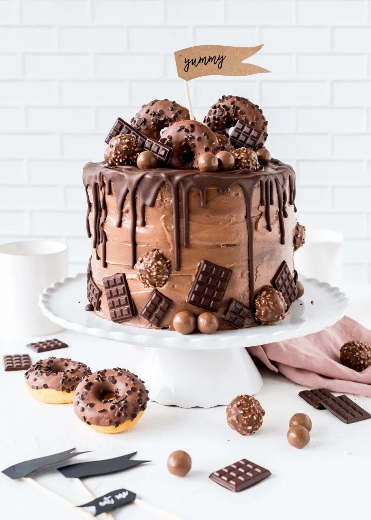 Fertig dekorierte Nutella Torte auf einem Tortenständer, umrahmt von Schokoladenstücken und Pralinen.