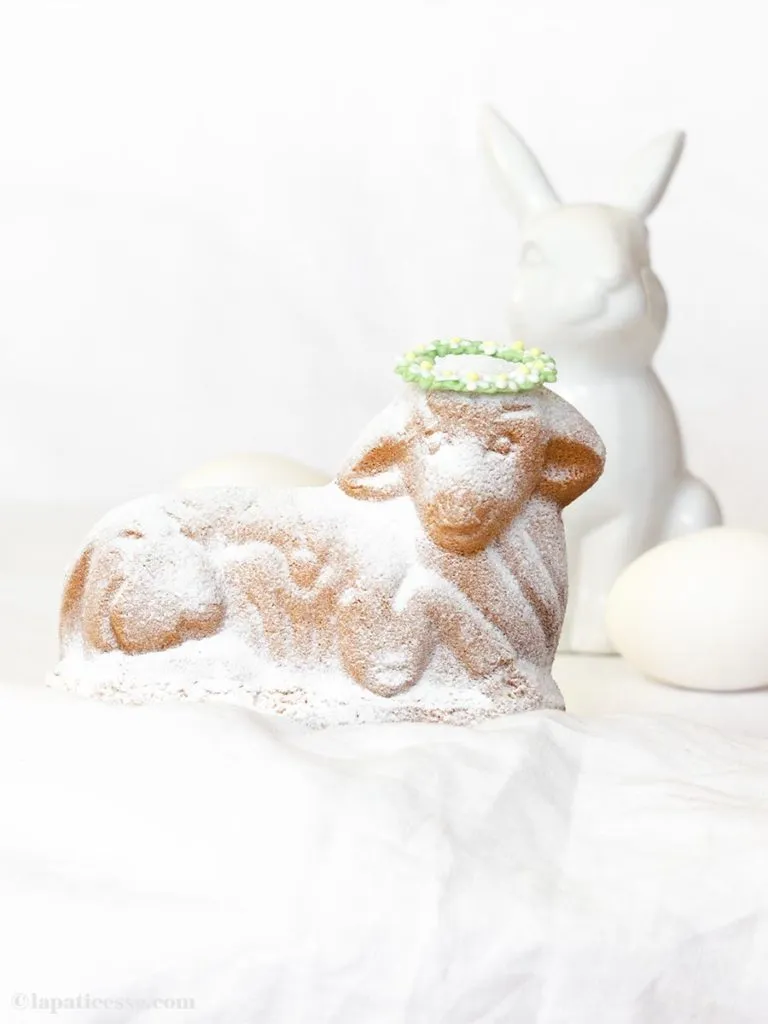 Fertig gebackenes und mit Puderzucker bestäubtes Osterlamm mit Blumenkranz, umgeben von Osterdekoration.