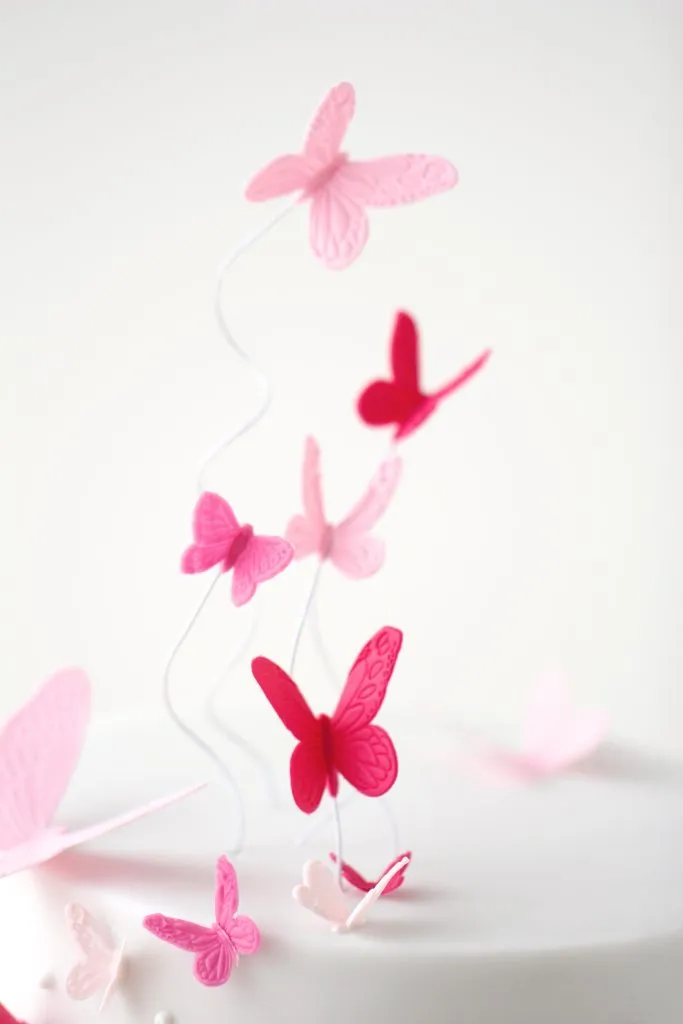 Filigrane Schmetterlinge auf gebogenen Blumendrähten, die über der Tauftorte schweben und ihr Tiefe verleihen