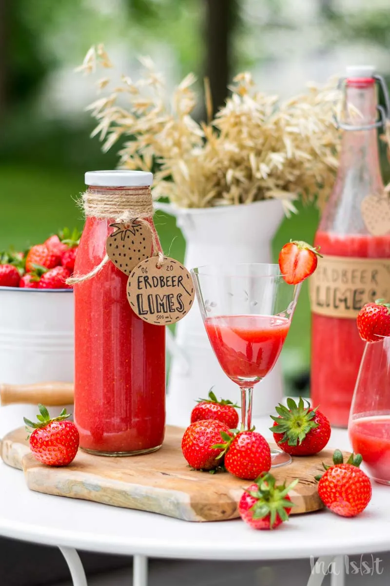 Frisch zubereiteter Erdbeerlimes in Glasflaschen, appetitlich angerichtet auf einem Gartentisch im Grünen für einen Sommerabend.