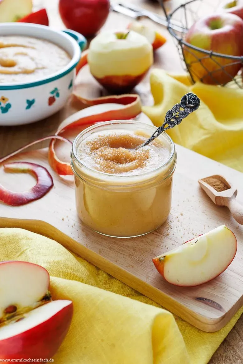 Frisch zubereitetes Apfelmus in einer Schale, daneben ein Löffel und ganze Äpfel, auf einem rustikalen Untergrund