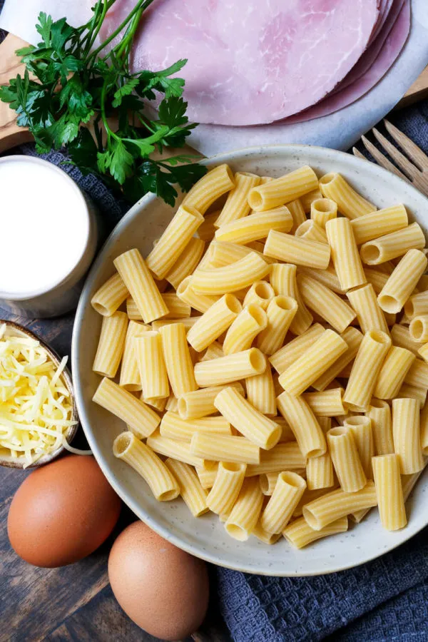 Frische Zutaten für einen Nudel-Schinken-Gratin: ungekochte Nudeln, gekochter Schinken, geriebener Käse, Sahne und Eier liegen auf einem Holztisch bereit