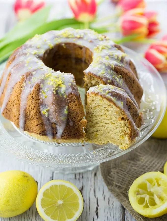 Frischer veganer Zitronenkuchen auf einer Glasplatte, umrahmt von frischen Zitronen und bunten Tulpen