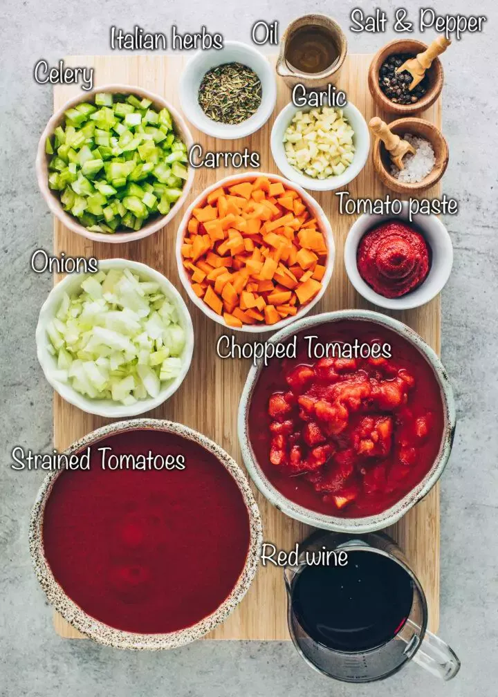 Frisches Gemüse und Gewürze wie Zwiebeln, Knoblauch, Olivenöl, Sellerie, Karotten, Tomaten, Tomatenmark, italienische Kräuter, Salz, Pfeffer und eine Flasche Rotwein auf einer Holzarbeitsfläche.