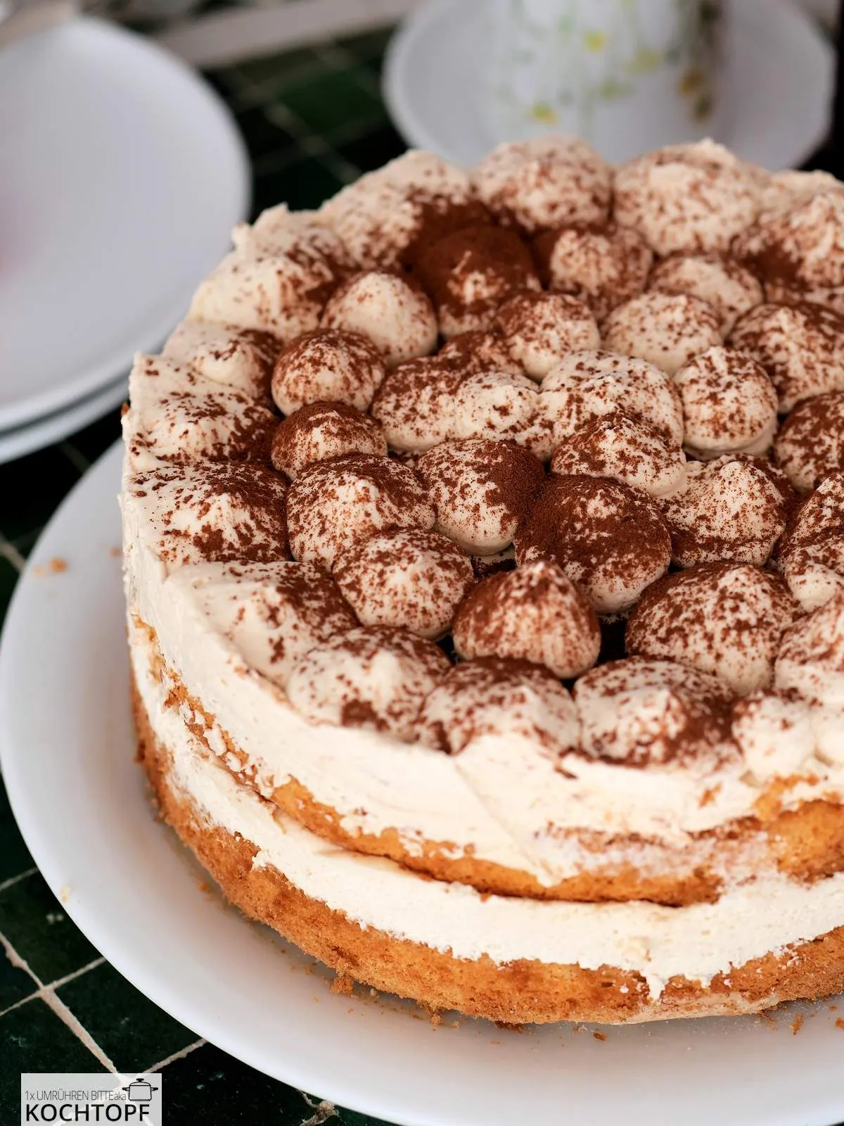 Ganze Tiramisu-Torte, fertig dekoriert mit Kakaopulver, präsentiert auf einem festlichen weissen Teller.