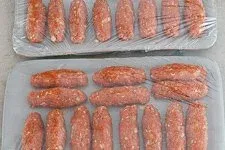 Geformte Cevapcici-Röllchen auf einem Teller, mit Frischhaltefolie abgedeckt zum Kühlen