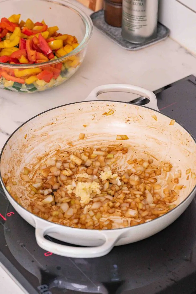 Gehackte Zwiebeln, die mit gehacktem Knoblauch in einer weißen Pfanne auf einem Herd sautiert werden, mit einer Schüssel geschnittener Paprika und Zucchini im Hintergrund.