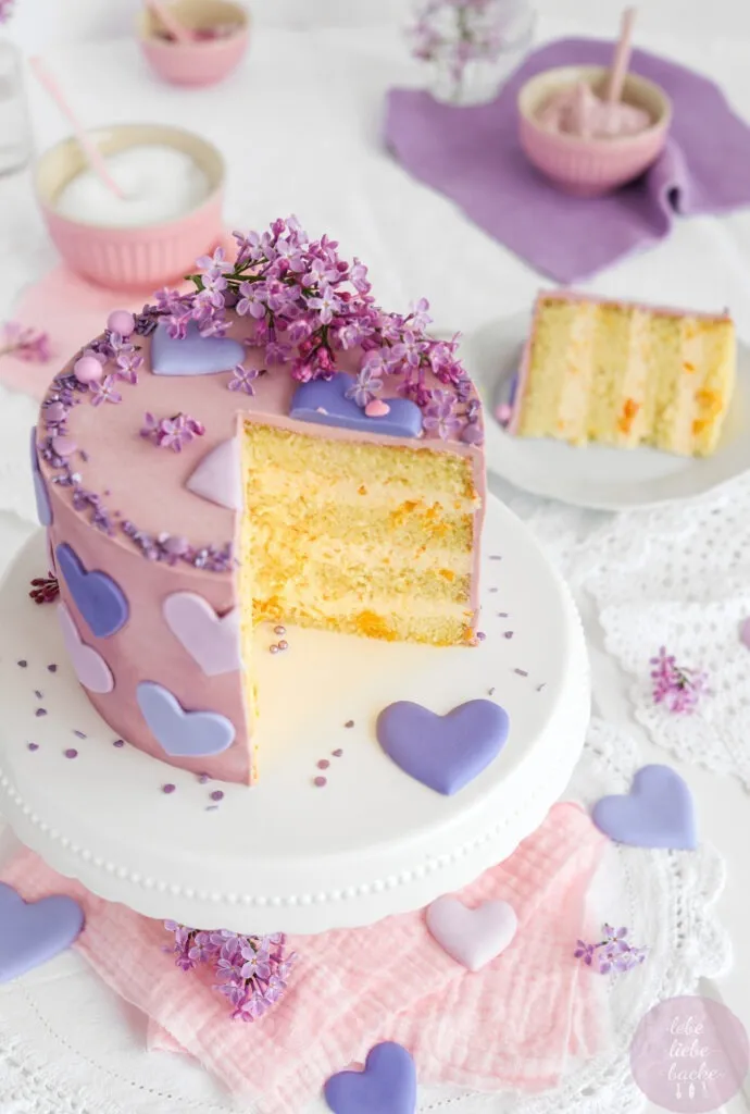 Gesamtaufnahme einer fertig dekorierten Käse-Sahne-Torte auf einem Tortenständer, umgeben von Flieder