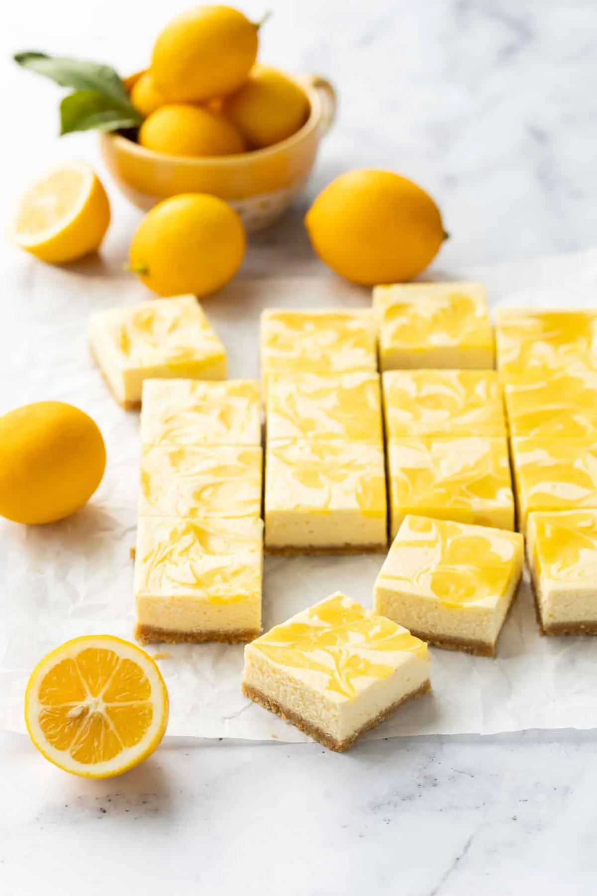 Geschnittene Meyer Zitronen-Käsekuchenriegel mit Lemon Curd Wirbeln auf Backpapier, dazu frische Zitronen.