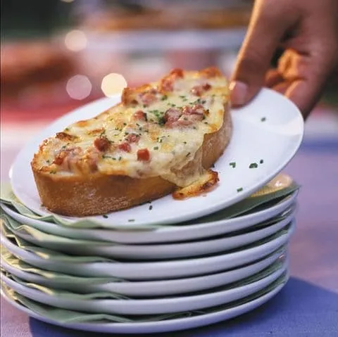 Goldbraun gratinierte Käse-Crostinis mit geschmolzenem Raclettekäse und Kräutern auf einem rustikalen Holzbrett.