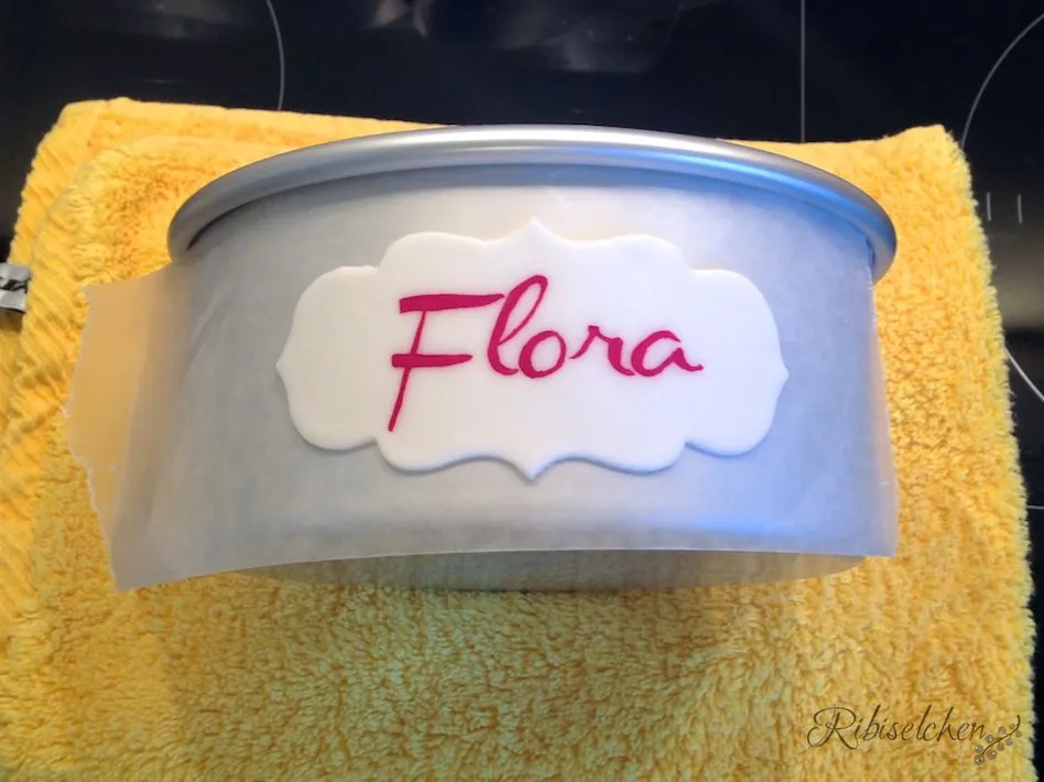 Handgemaltes Namensschild &quot;Flora&quot; auf einer Fondant-Plaque, verziert mit einem pinken Fondantrand