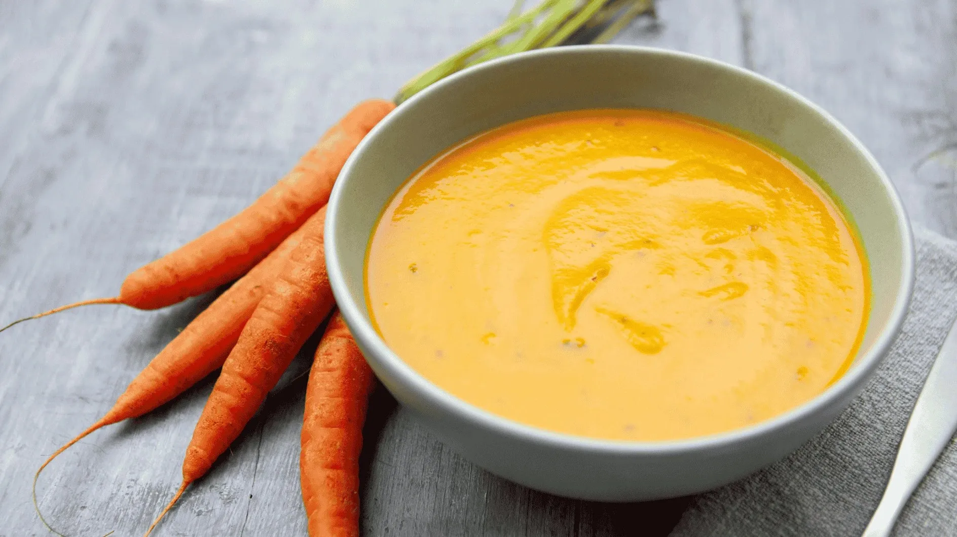 Hausgemachte Karottensuppe in einer grünen Schüssel, mit einem Bund Karotten daneben, auf groben Holztisch