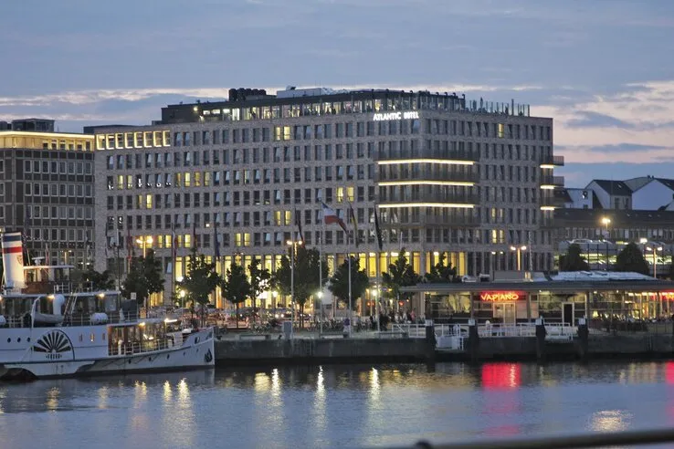 Hotelgebäude an der Uferpromenade in der Dämmerung, beleuchtet, mit Flaggen und Restaurant im Vordergrund.