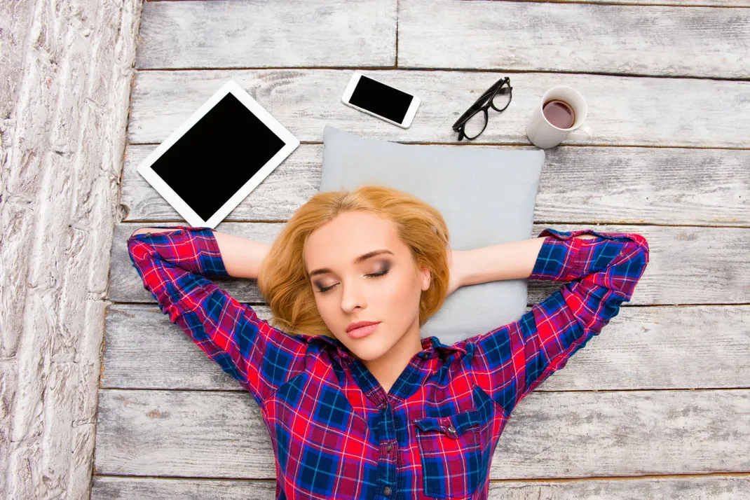 Junge Frau mit blonden Haaren liegt entspannt auf einem Kissen auf einem Holzfußboden, umgeben von Tablet, Smartphone, Brille und einer Tasse Kaffee.