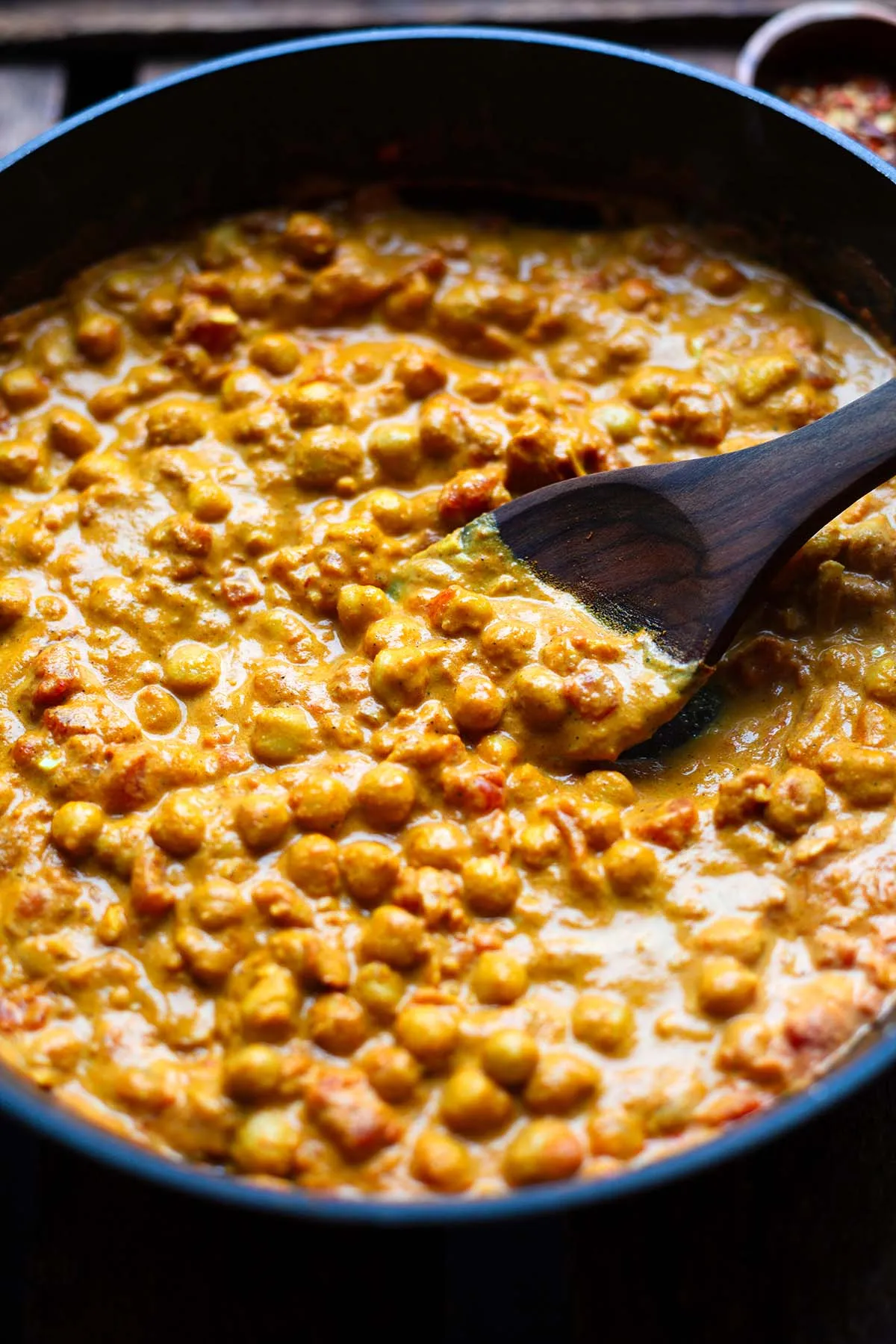 Kichererbsen-Curry mit Kokosmilch in einer Pfanne, während es köchelt, mit einem Holzlöffel. Zeigt die Zubereitung eines veganen Kichererbsen Rezepts.