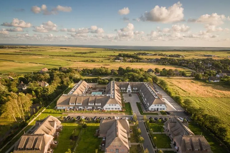 Luftaufnahme eines Hotels mit Innenhof, umgeben von grünen Feldern und vereinzelten Gebäuden unter blauem Himmel.
