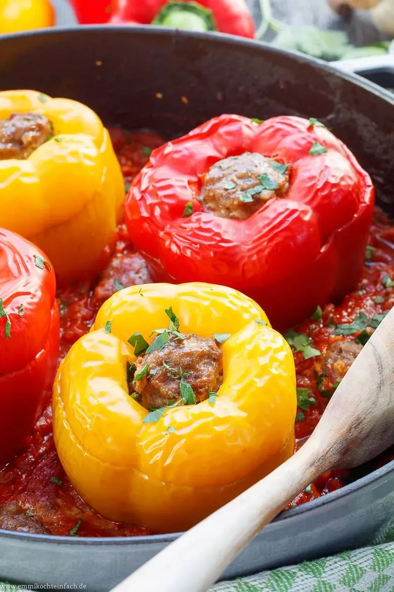 Mehrere bunte gefüllte Paprikaschoten mit Hackfleisch und Deckel in einer Auflaufform, umgeben von reichlich Tomatensoße.