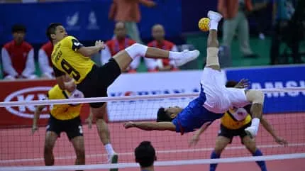 Mehrere Männer in Sportkleidung stehen auf einem Feld, das durch ein Netz getrennt wird. Zwei Männer direkt am Netz befinden sich im Sprung und strecken den Fuß nach dem Ball aus, der sich in der Luft befindet.