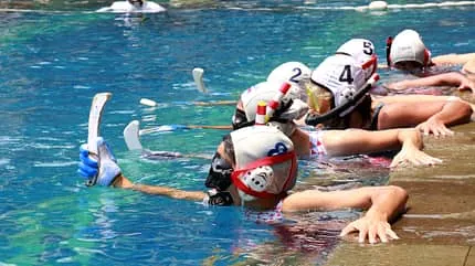 Mehrere Schwimmer:innen in Badebekleidung halten sich mit einer Hand am Rand eines Schwimmbeckens fest. In der anderen Hand halten sie einen kurzen Schläger.