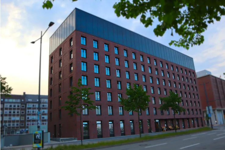 Modernes Hotelgebäude aus rotem Backstein mit großen Fenstern, umgeben von Bäumen und der Straße im Vordergrund.