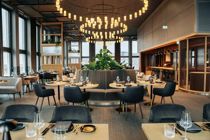Modernes Hotelrestaurant mit eleganten Tischen, schwarzen Stühlen, großen Fenstern und stilvollen Kronleuchtern.