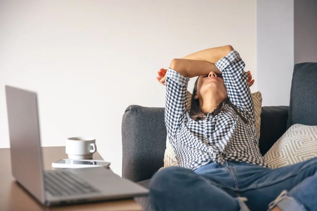 Müde Frau lehnt sich mit verschränkten Armen auf Sofa, Laptop und Smartphone liegen vor ihr.