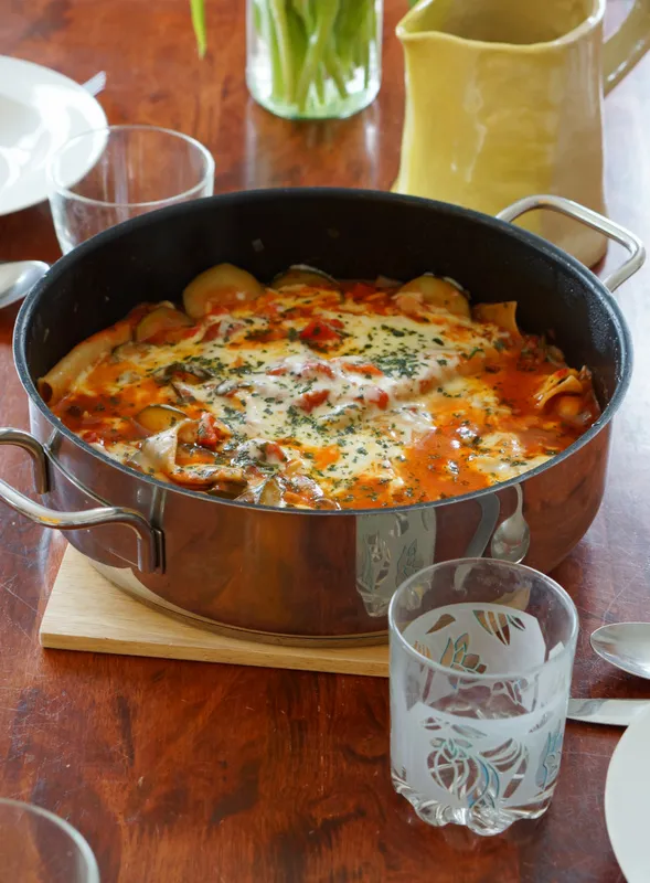 Nahaufnahme einer dampfenden Portion Gemüse Lasagne aus der Pfanne, garniert mit frischem Basilikum und Parmesan, die auf einem rustikalen Holztisch serviert wird. Der Fokus liegt auf der Textur der Nudeln, des Gemüses und des geschmolzenen Käses.