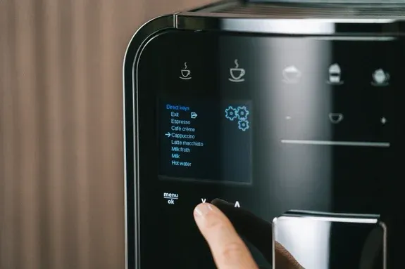 Nahaufnahme einer Hand, die auf einem Kaffeevollautomat mit Milchsystem eine Kaffeespezialität auswählt, typisch für den modernen deutschen Kaffeegenuss.