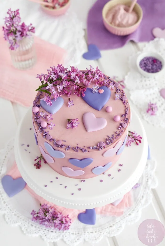 Nahaufnahme einer Käse-Sahne-Torte mit lila und rosa Buttercreme-Tupfen und frischen Fliederblüten als Geburtstagsdeko