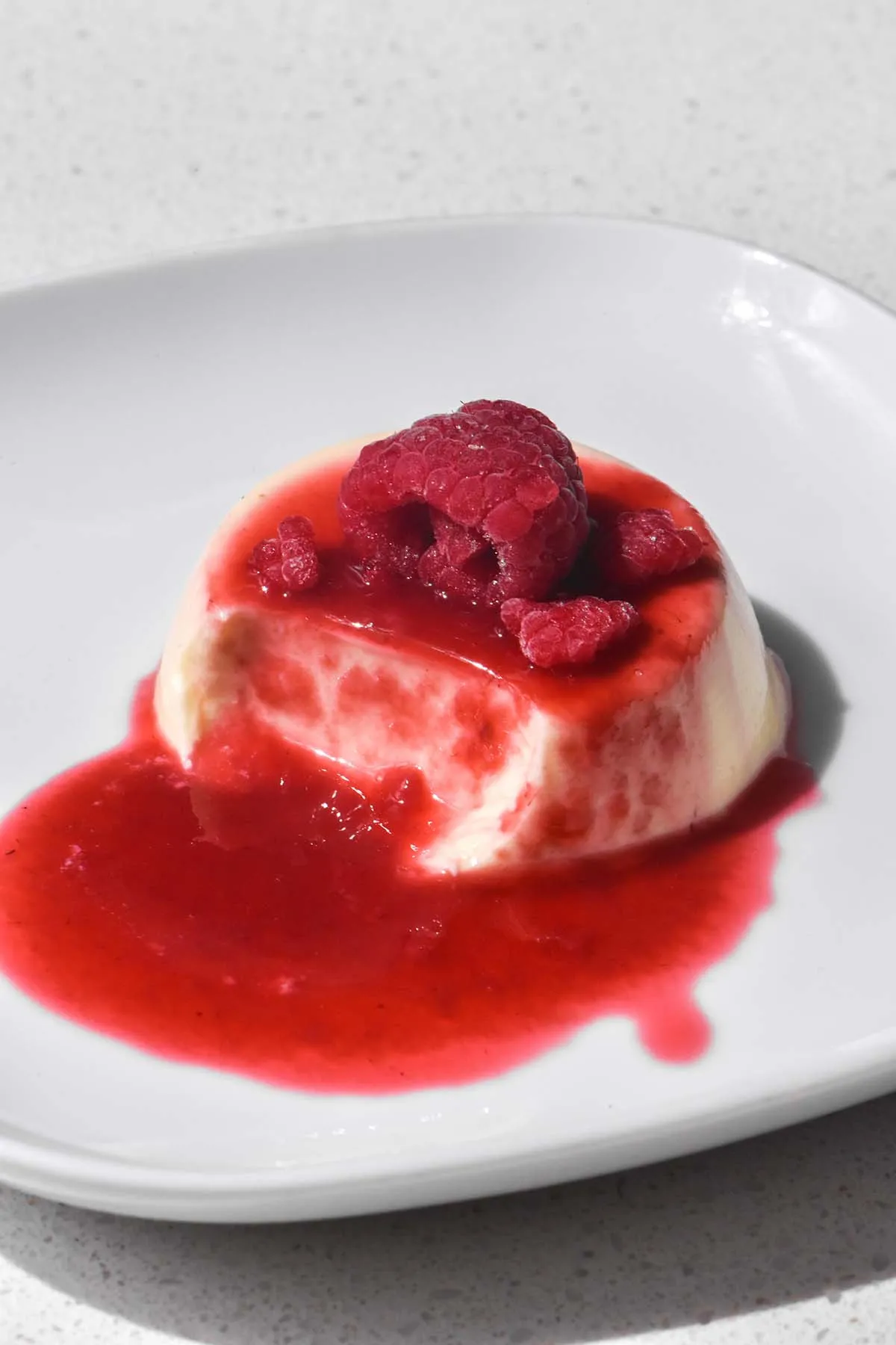 Nahaufnahme einer Panna Cotta mit Himbeercoulis und frischen Himbeeren. Ein Löffel entnimmt einen Teil, der die cremige Textur zeigt.