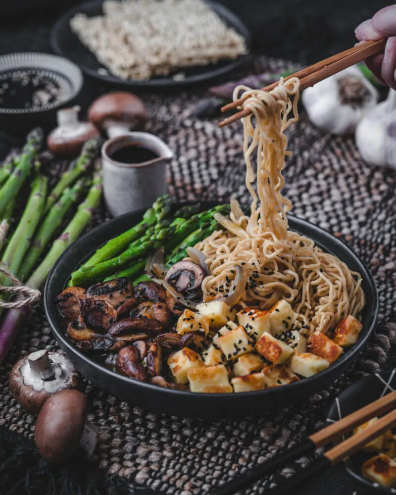 Nahaufnahme einer vegetarischen Ramen Suppe in einer rustikalen Schale, aufgewickelte Nudeln werden mit Essstäbchen gehalten, umrahmt von Halloumi, Spargel und Pilzen