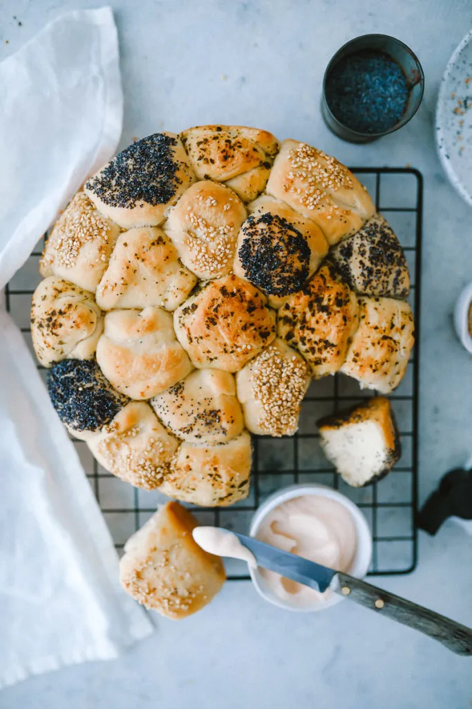 Nahaufnahme eines Brötchenkranzes, dekoriert mit Mohn und Sesam, umgeben von kleinen Schälchen mit Dips auf einem Holztisch.