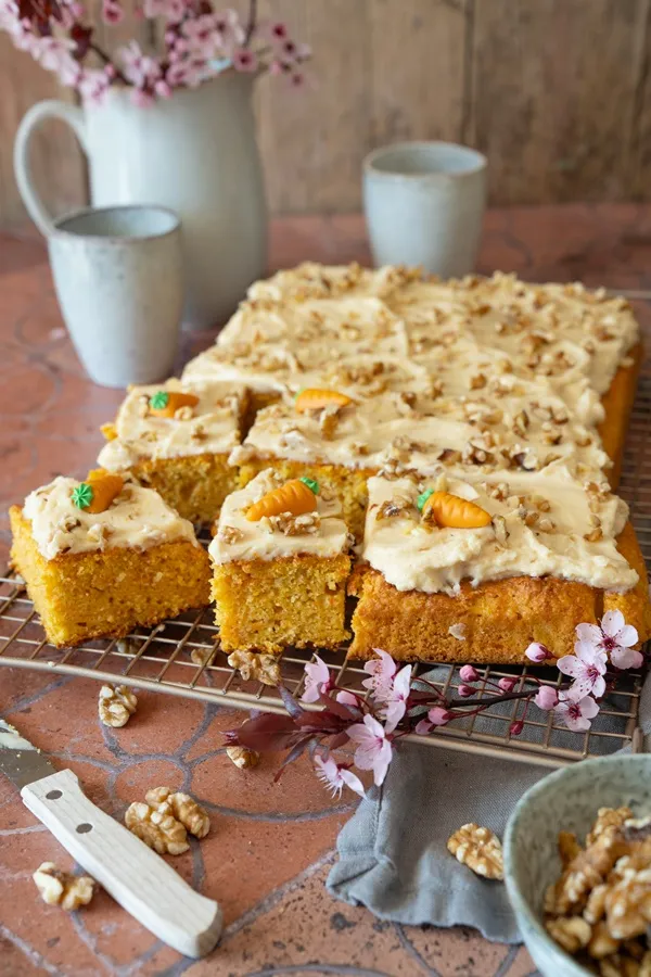 Nahaufnahme eines Stücks saftigen Rüblikuchens vom Blech, reichlich bedeckt mit weißem Frischkäse-Frosting und Marzipanmöhren.