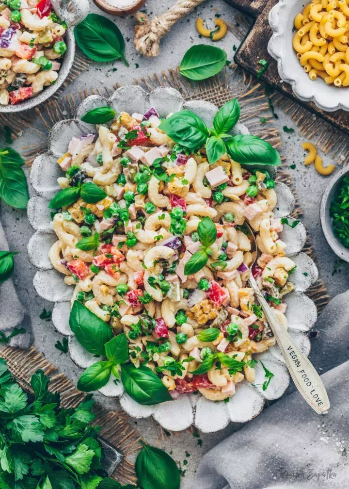Nahaufnahme eines veganen Nudelsalats mit Spiralnudeln, Erbsen, Mais, Paprika, Gurken und einer cremigen, hellen Sauce, garniert mit frischen Kräutern. Die Komposition betont die frischen Zutaten und die appetitliche Textur des Salats.