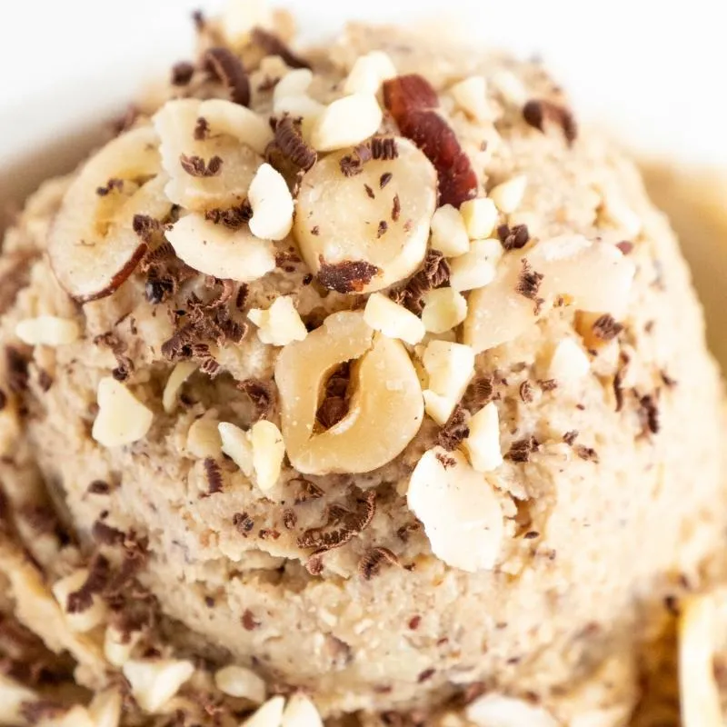 Nahaufnahme von hausgemachtem Keto Haselnusseis in einer Glasschale, garniert mit gehackten Haselnüssen und einem Minzblatt. Das Eis hat eine cremige Textur und eine appetitliche Farbe. Alt-Text: Cremiges Keto Haselnusseis Rezept - Genuss ohne Kohlenhydrate