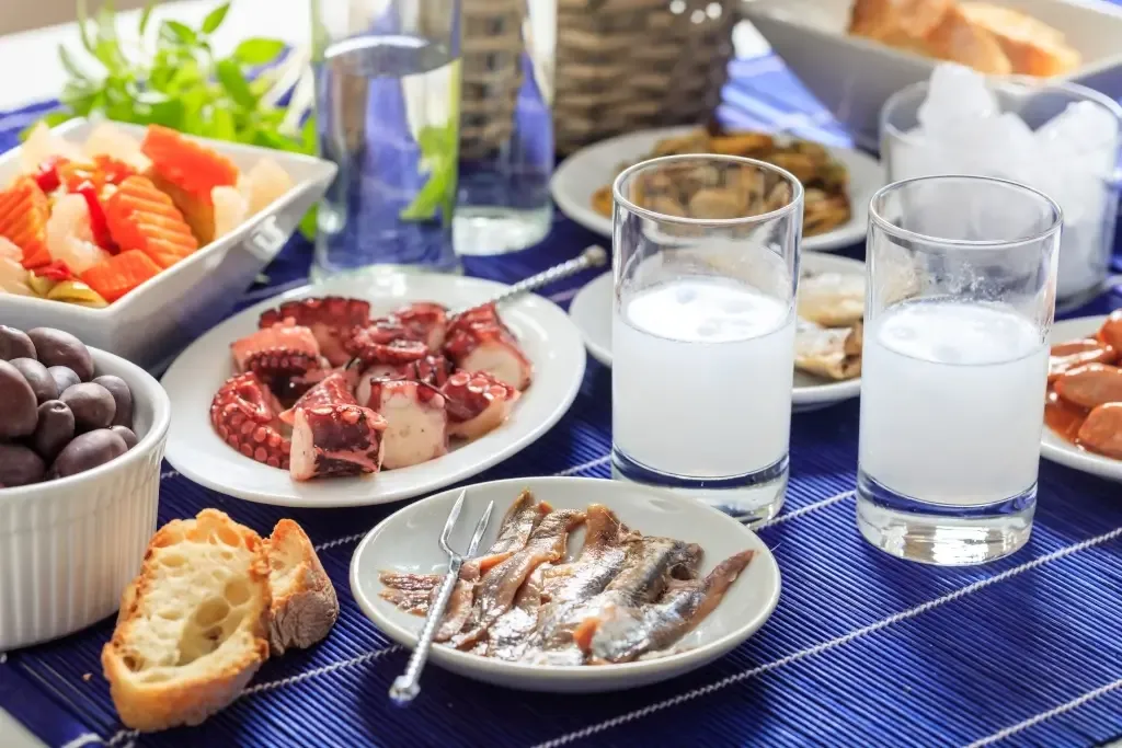 Ouzo-Glas mit milchiger Trübung, serviert mit einer Auswahl traditioneller griechischer Mezedes (Vorspeisen) auf einem Holztisch, perfekt zum Genuss des Ouzo Getränk.