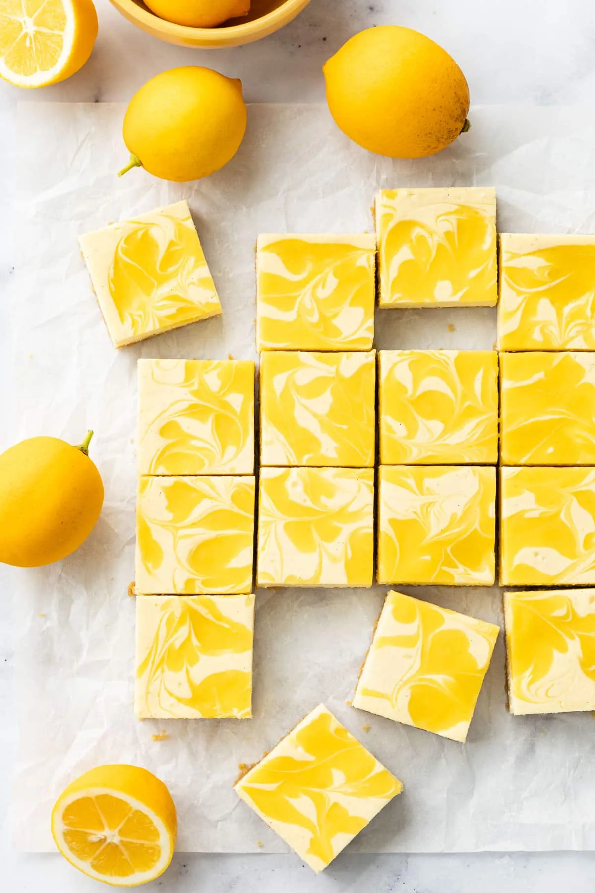 Quadrate von Meyer Zitronen-Käsekuchenriegeln mit leuchtend gelben Lemon Curd Wirbeln auf Backpapier, umgeben von frischen Zitronen.